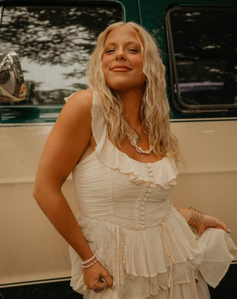 Country star Ashley Anne, with long blonde hair, smiles confidently in a ruffled white dress in front of a green and white vehicle, accessorized with layered necklaces, bracelets, and rings.