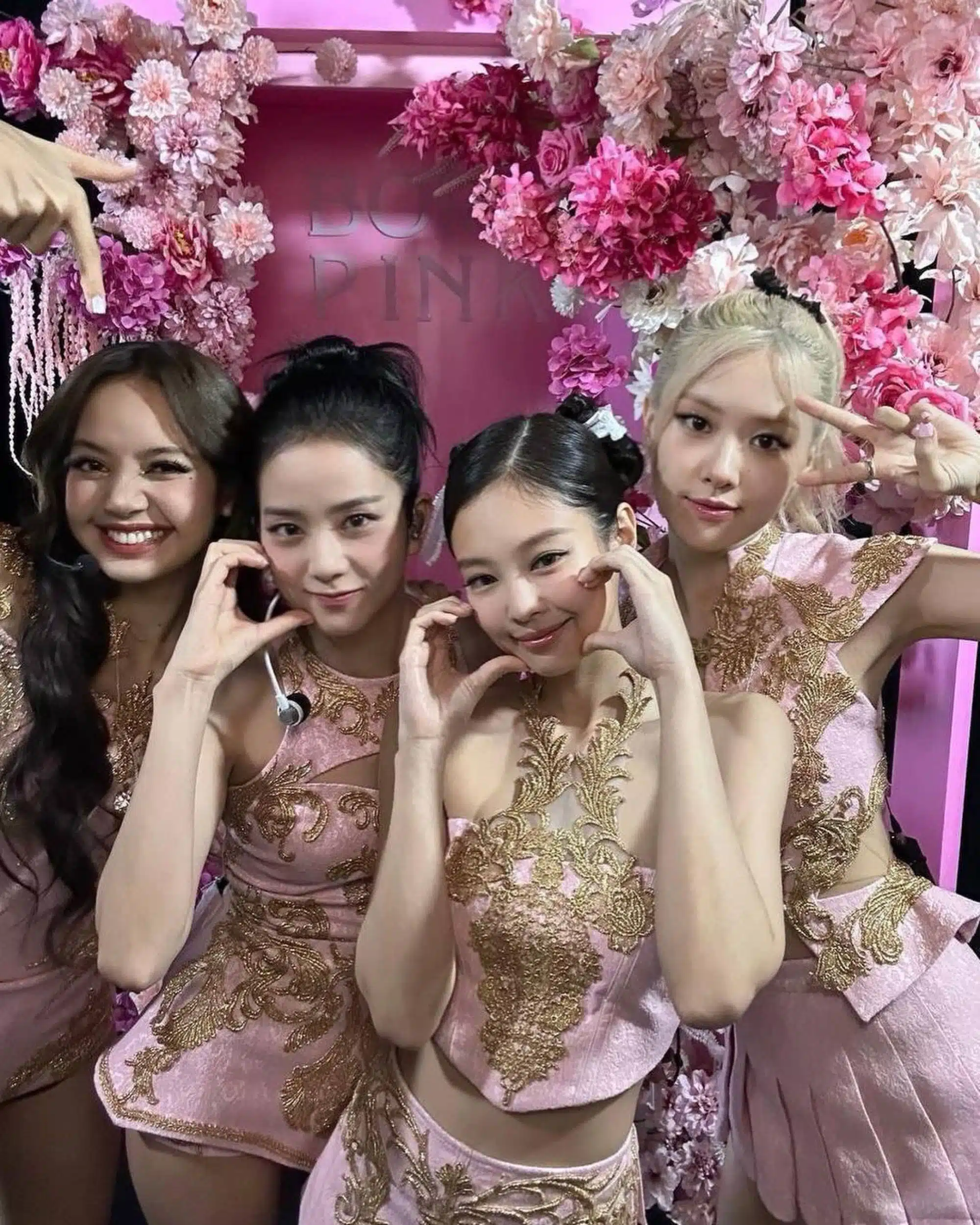 Four women, dressed in matching pink and gold outfits, pose together in front of a pink floral backdrop, making heart shapes with their hands and smiling at the camera, channeling BLACKPINKs style as they prep for the DEADLINE world tour.