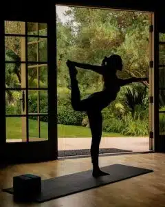 A person practices a standing yoga pose indoors on a mat, silhouetted against open double doors that reveal a lush, green garden outside—an inviting scene to refresh and relax, reminiscent of the tranquility at Cal-a-Vie Health Spa.