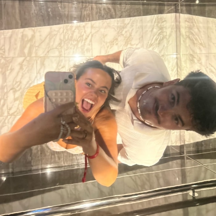 A woman and man smile up at a mirrored ceiling, taking a selfie. The woman is holding the phone, smiling widely, while the man stands next to her, looking at the camera. Marble flooring is visible in the reflection.