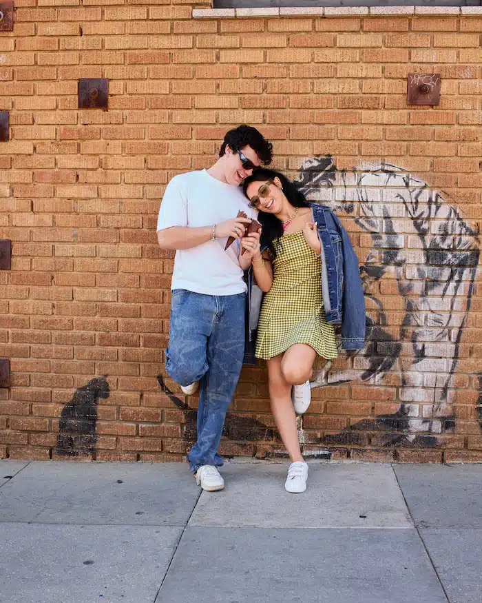 Jentzen and Elliana lean against a graffiti-covered brick wall, smiling as they look at a phone together. The woman in a yellow dress and jean jacket, the man in white shirt and jeans—these relationship heroes look relaxed and happy.