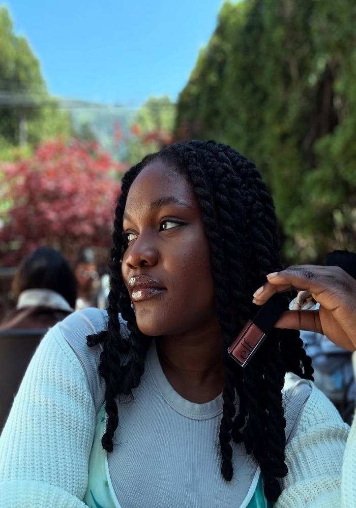 A woman with long braided hair sits outdoors, holding a trending tube of lip gloss near her face. She is wearing a light-colored sweater and looking to the side, with trees and red foliage in the background.