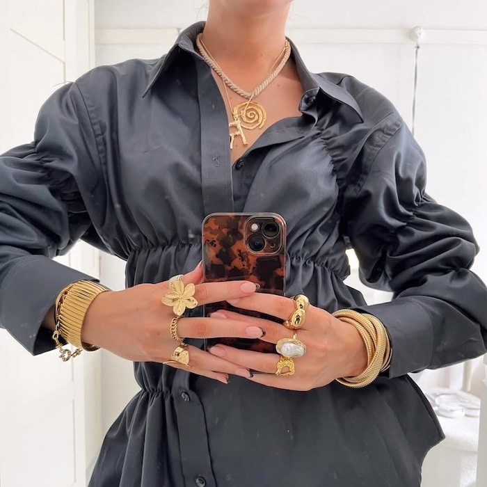 A person wearing a dark, long-sleeved shirt holds a phone with both hands, displaying multiple gold rings, bracelets, and a necklace with a spiral pendant. The background is a bright, white room.