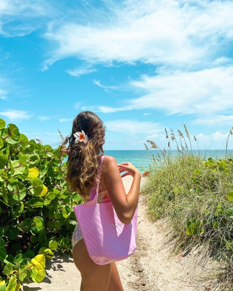 A woman with wavy hair and a white flower behind her ear stands on a sandy path toward the ocean, carrying a pink beach bag, surrounded by green foliage under a bright blue sky with scattered clouds.