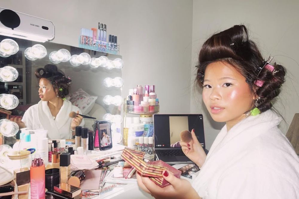 A woman in a white robe with hair rollers applies makeup at a vanity filled with cosmetics, facing a brightly lit mirror. Her reflection and open laptop are visible on the counter.