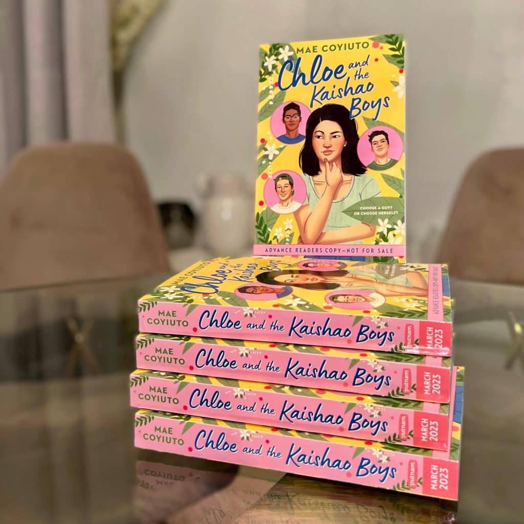 A stack of five books, Chloe and the Kaishao Boys by Mae Coyiuto, is arranged on a table with one standing upright. The colorful cover, perfect for fans of TSITP or similar reads, features illustrated characters and floral accents.