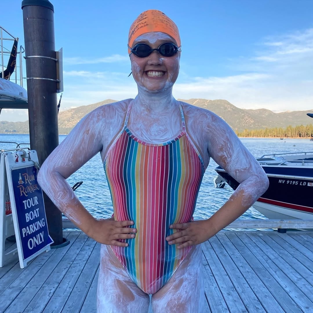 A smiling swimmer wearing goggles, an orange swim cap, and a colorful striped swimsuit stands on a wooden dock with a lake and mountains in the background. Her skin is covered in a thick layer of white sunscreen.
