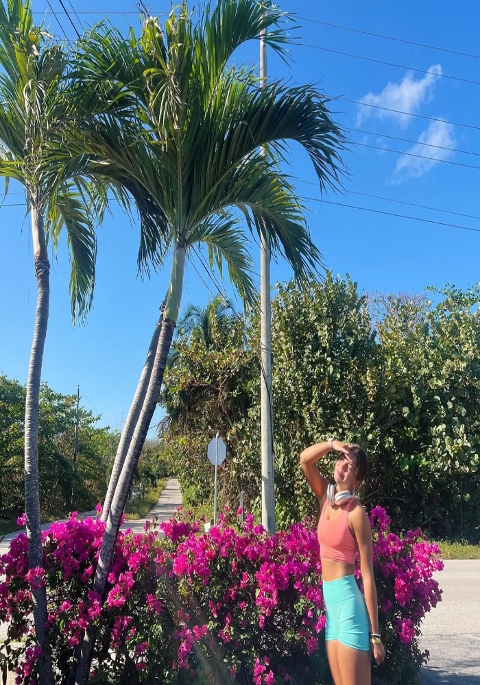 A woman in athletic wear stands among bright pink flowers, shielding her eyes from the sun. Palm trees and a clear blue sky set the scene for a summer fitness moment, capturing the spirit of a fun workout in a tropical paradise.