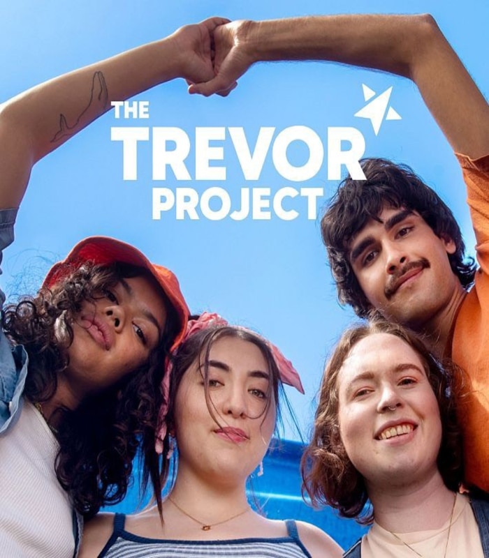 Four young people stand close together, smiling and holding hands above their heads under a bright blue sky. The Trevor Project logo appears above them in white text with a paper airplane icon, representing LGBTQ+ crisis support.