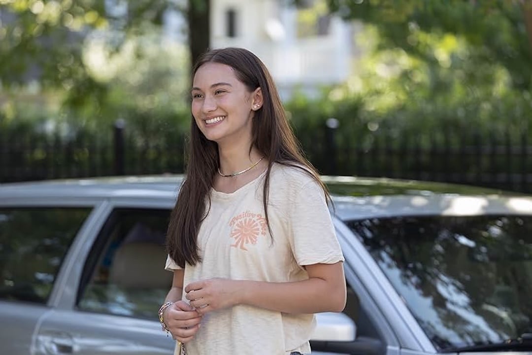 A young woman with long brown hair smiles while standing outside in front of a parked silver car, showcasing summer fashion inspired by The Summer I Turned Pretty. She wears a light-colored t-shirt, bracelets, with trees and a fence in the background.