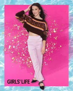 A smiling young woman poses against a vibrant pink and gold background. She wears a patterned brown sweater, light pink pants, and black shoes. The words “GIRLS’ LIFE” appear in the bottom left corner, celebrating the 2024 Barbizon Model Search contest winner.