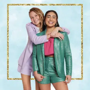 Two smiling young women pose together, dressed in vibrant suits perfect for a summer vision board. One wears a sparkly green suit with shorts; the other hugs her from behind in a purple blazer. A light blue backdrop with a gold glitter border completes the scene.