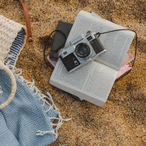 A vintage camera rests on an open book placed on sandy ground, next to a blue and white patterned blanket and a woven bag. The scene suggests a relaxing day at the beach with photography, reading, and catching up on your summer TBR.
