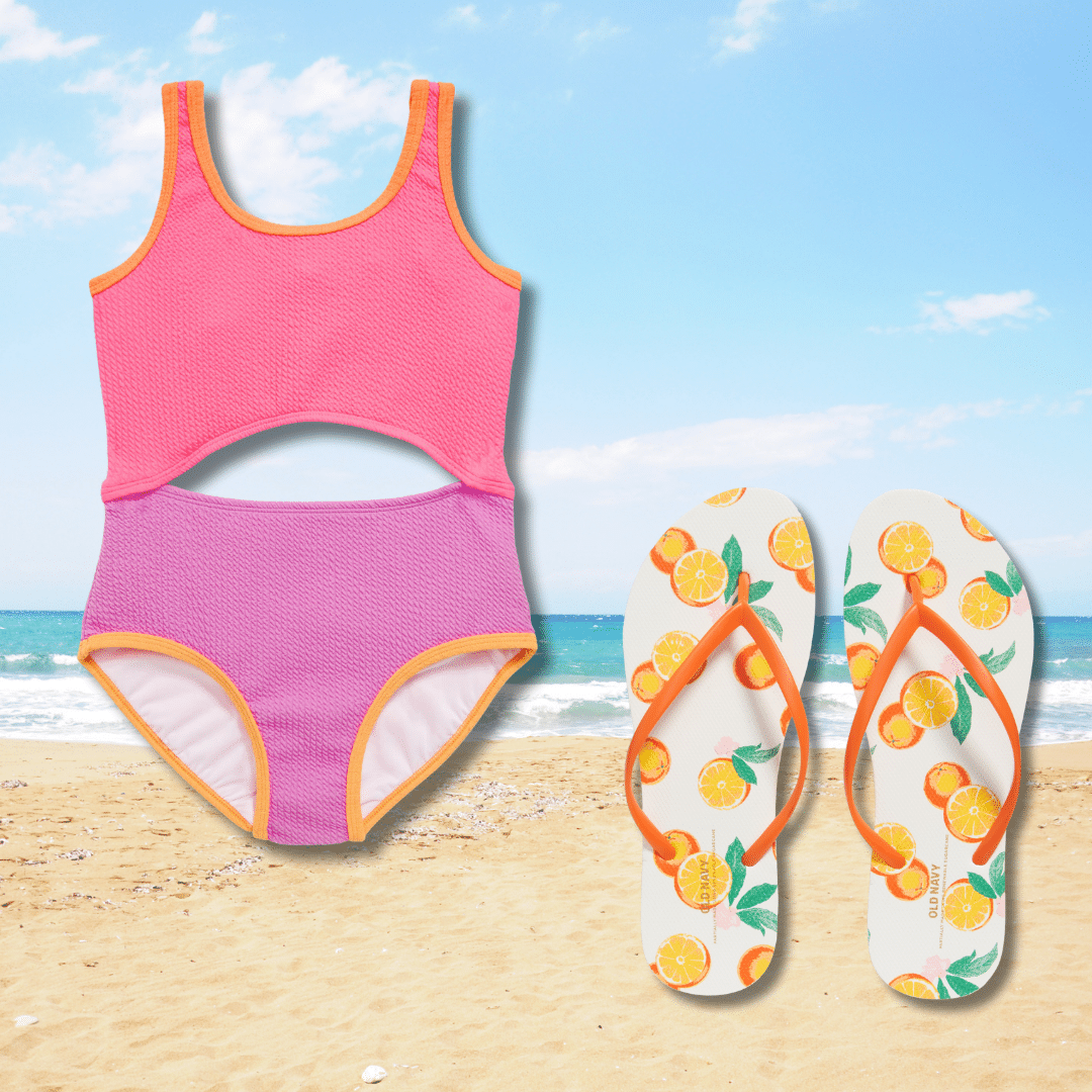 A pink and purple cutout swimsuit and a pair of white flip-flops with orange fruit patterns are placed on a sandy beach, perfect for sunny day staycations or updating your summer swimwear. The ocean and blue sky complete the scene.
